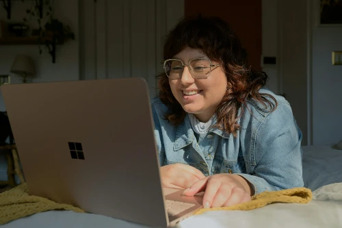 Ung kvinna söker jobb på sin laptop, liggandes på mage i sängen